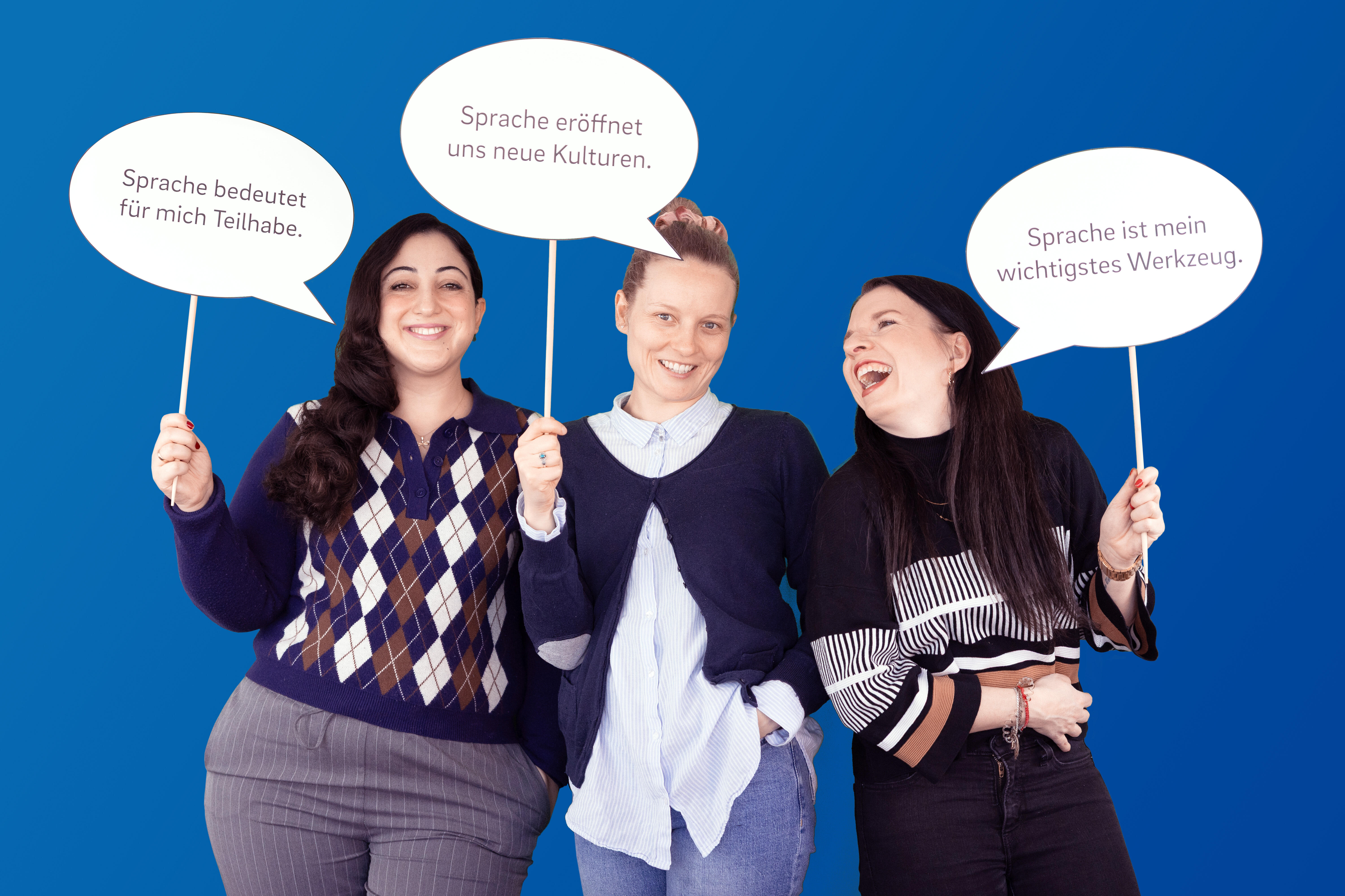 Drei fröhliche Mitarbeitende der VDV-Akademie halten Schilder hoch, auf denen Vorteile von Sprache im Berufskontext stehen.