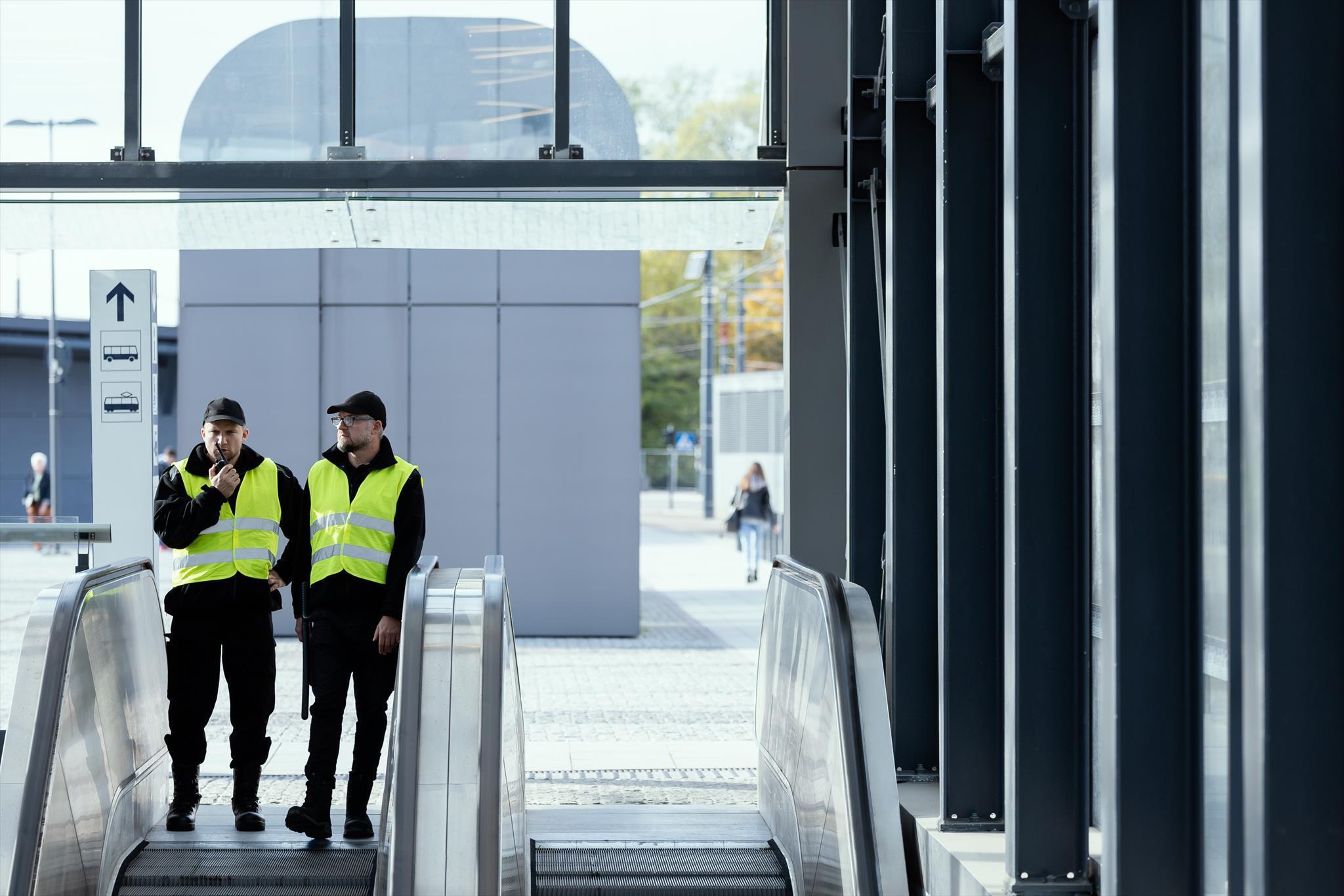 Zwei Männer in gelben Warnwesten und dunkler Kleidung betreten die Rolltreppe eines Bahnhofs.
