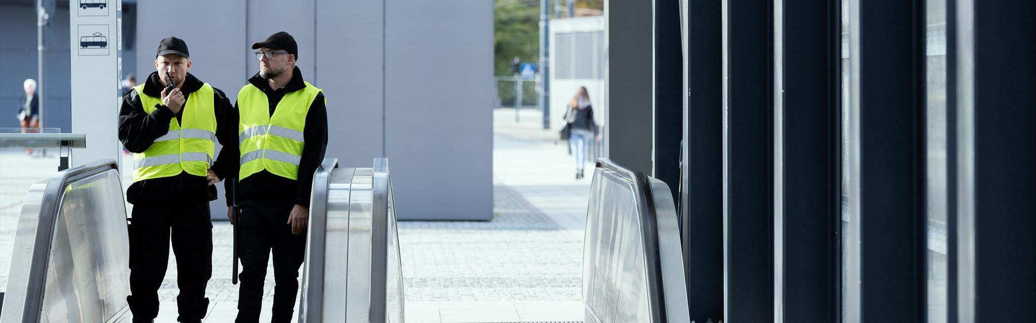 Zwei Männer in gelben Warnwesten und dunkler Kleidung betreten die Rolltreppe eines Bahnhofs.