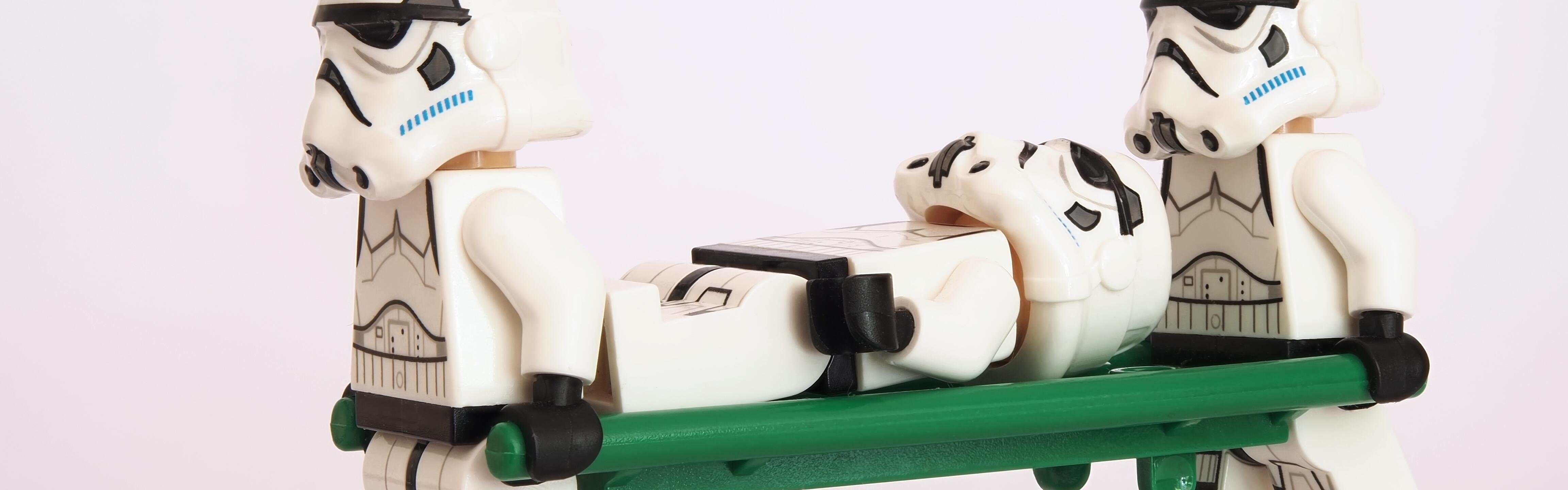 Zwei Legofiguren, die aussehen wie Stormtrooper, tragen als Sanitäter eine andere Stormtrooper-Legofigur auf einer Trage weg.