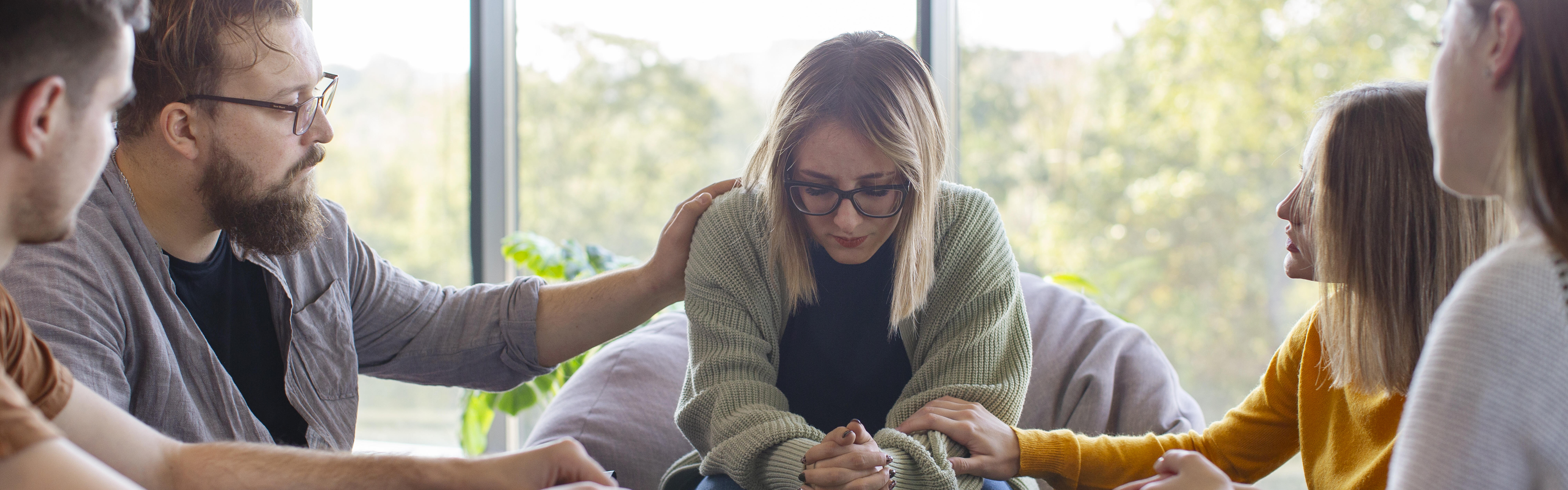 Fünf Personen sitzen im Kreis um eine Frau und zeigen ihr Mitgefühl, indem sie ihre Schulter und ihre Arme berühren.