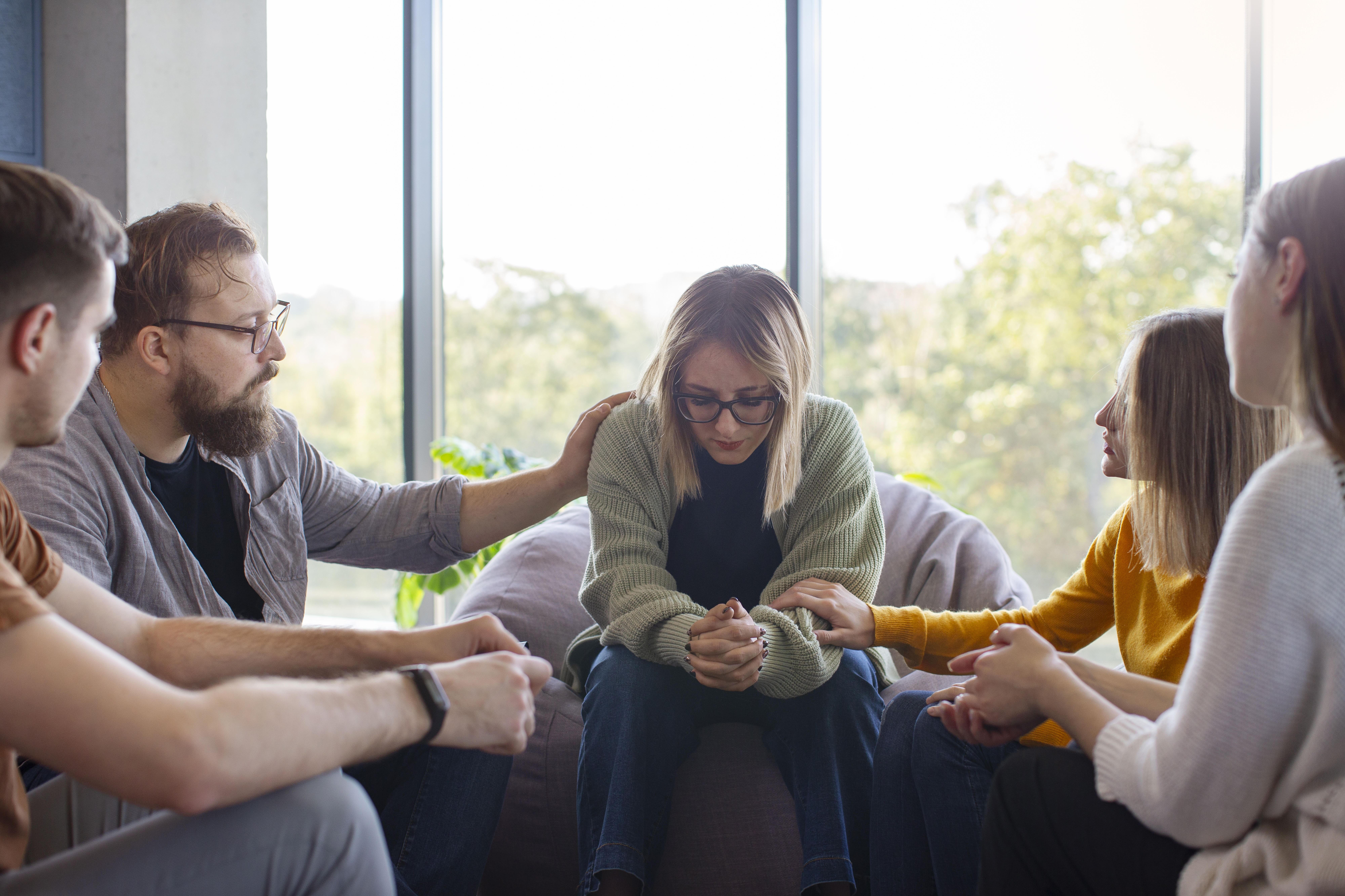 Fünf Personen sitzen im Kreis um eine Frau und zeigen ihr Mitgefühl, indem sie ihre Schulter und ihre Arme berühren.