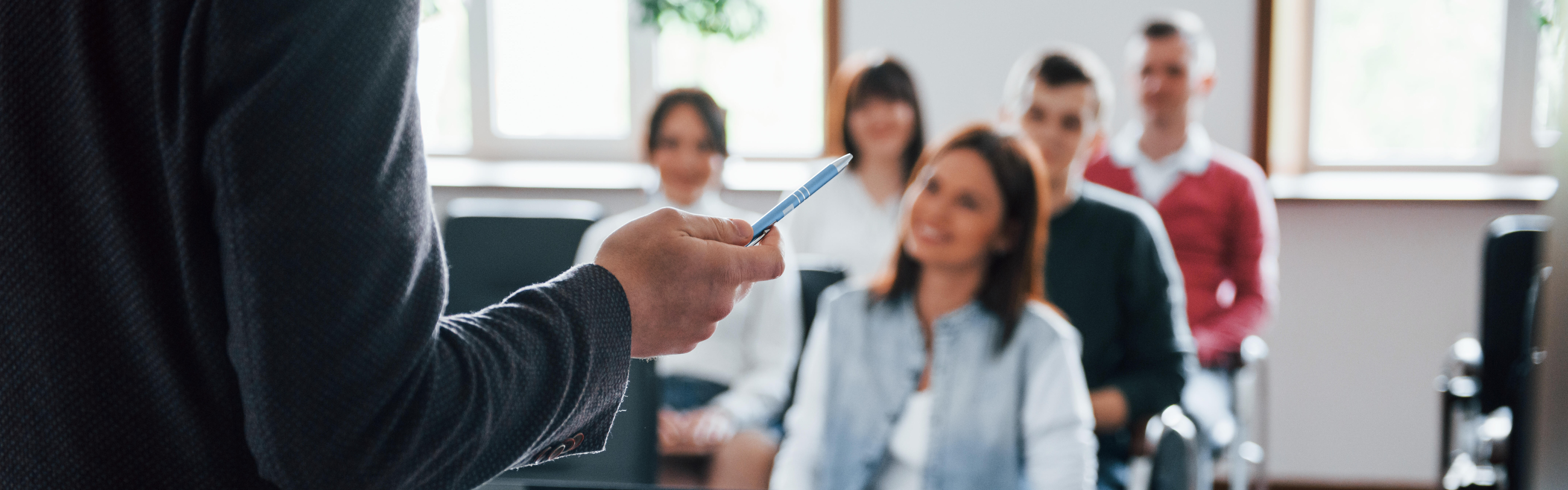 Eine Gruppe sitzt in einem Raum und hört einer Person zu, deren Rückenansicht man vor einem Laptop sieht.