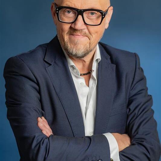 Auf dem Bild ist der Dozent Jens Blaufuss zu sehen. Er trägt eine markante, dunkle Brille. Sein Hemd ist oben leger geöffnet und er hat ein dunkels Sakko an. Mit verschränkten Armen beugt er sich lächeln zur Kamera.