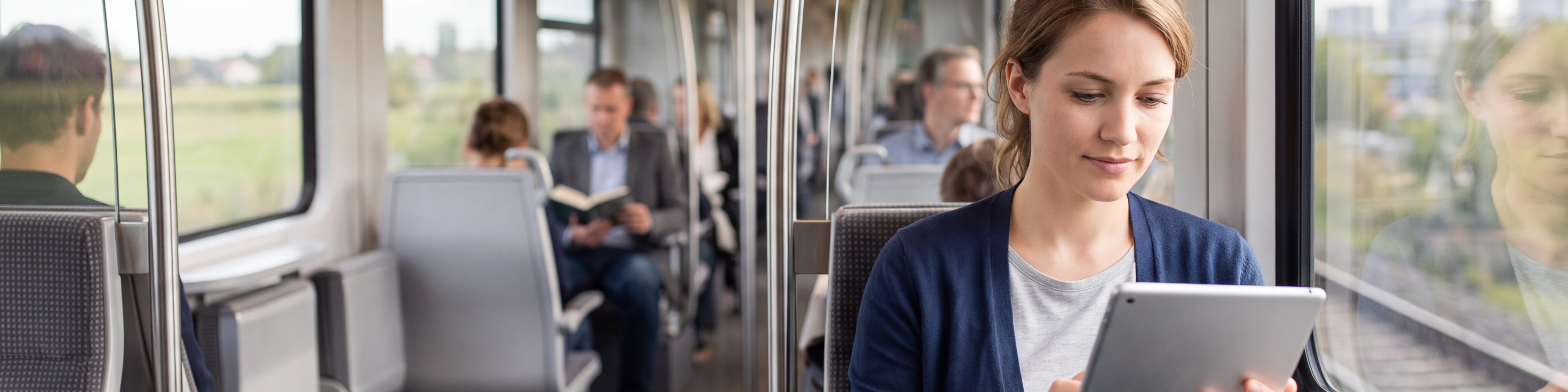Eine Frau sitzt in einem modernen Zug am Fenster und bedient ein Tablet. Sie trägt einen dunkelblauen Cardigan und ein graues Oberteil. Im Zug sitzen weitere Fahrgäste, einige lesen oder arbeiten. Durch die großen Fenster sind eine Landschaft und Gebäude zu sehen