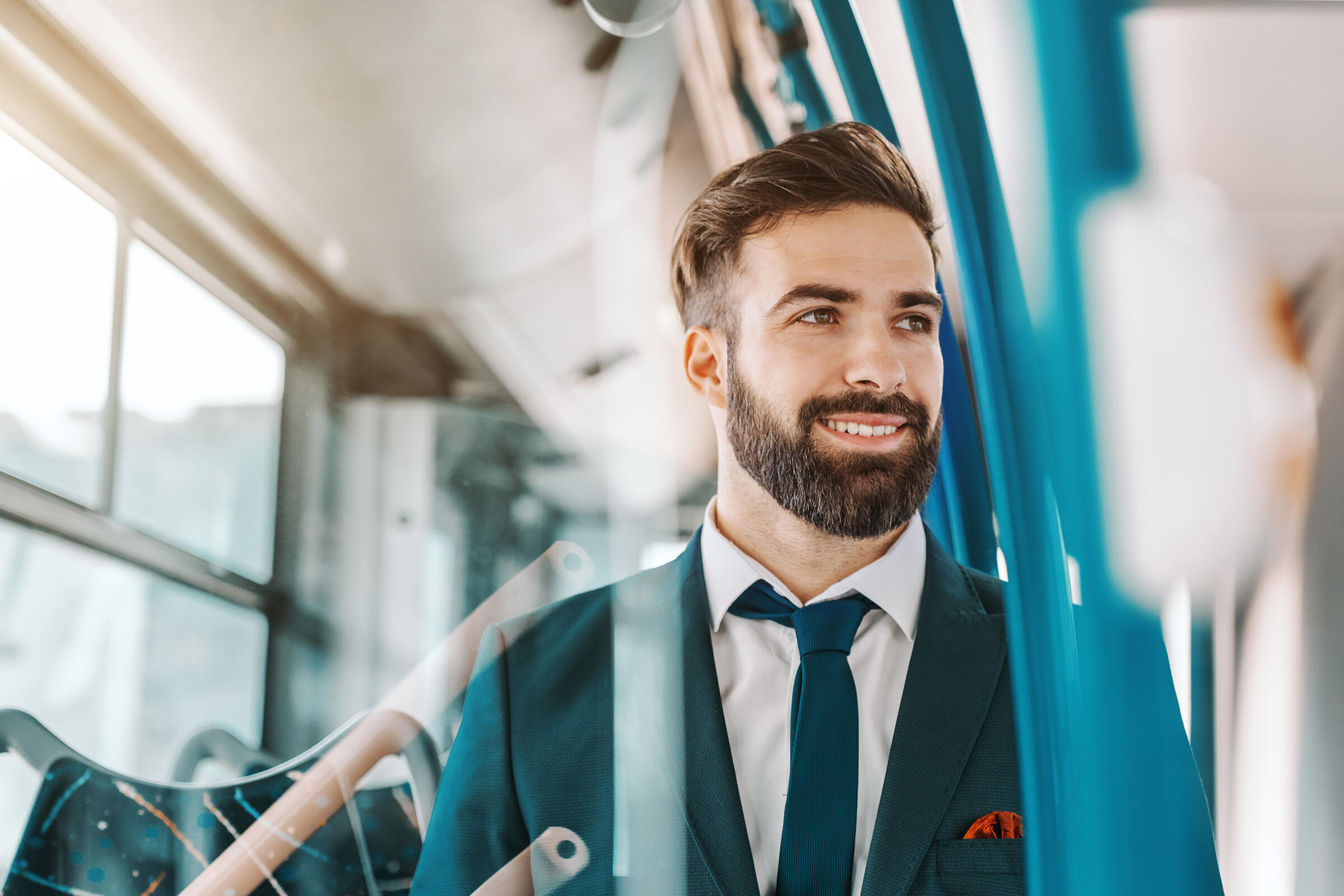 Nahaufnahme eines lächelnden bärtigen Geschäftsmannes in formeller Kleidung, der in einem öffentlichen Verkehrsmittel sitzt und aus dem Fenster schaut.
