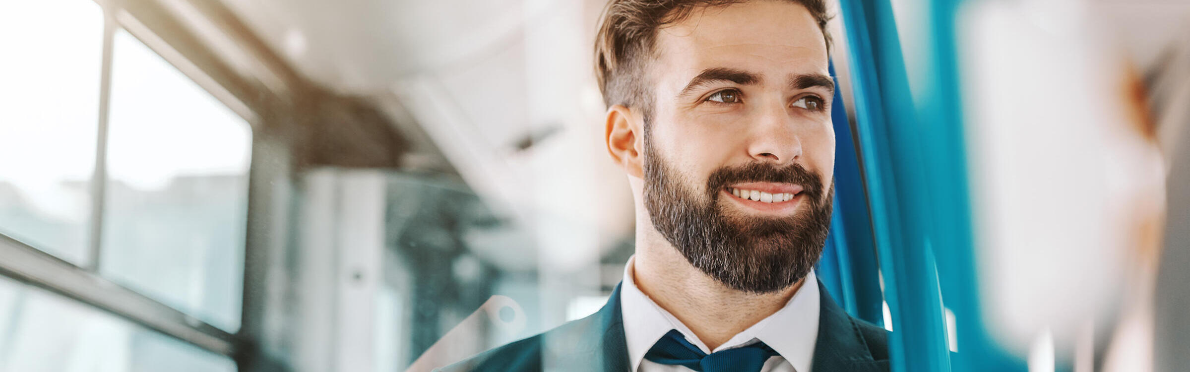 Nahaufnahme eines lächelnden bärtigen Geschäftsmannes in formeller Kleidung, der in einem öffentlichen Verkehrsmittel sitzt und aus dem Fenster schaut.