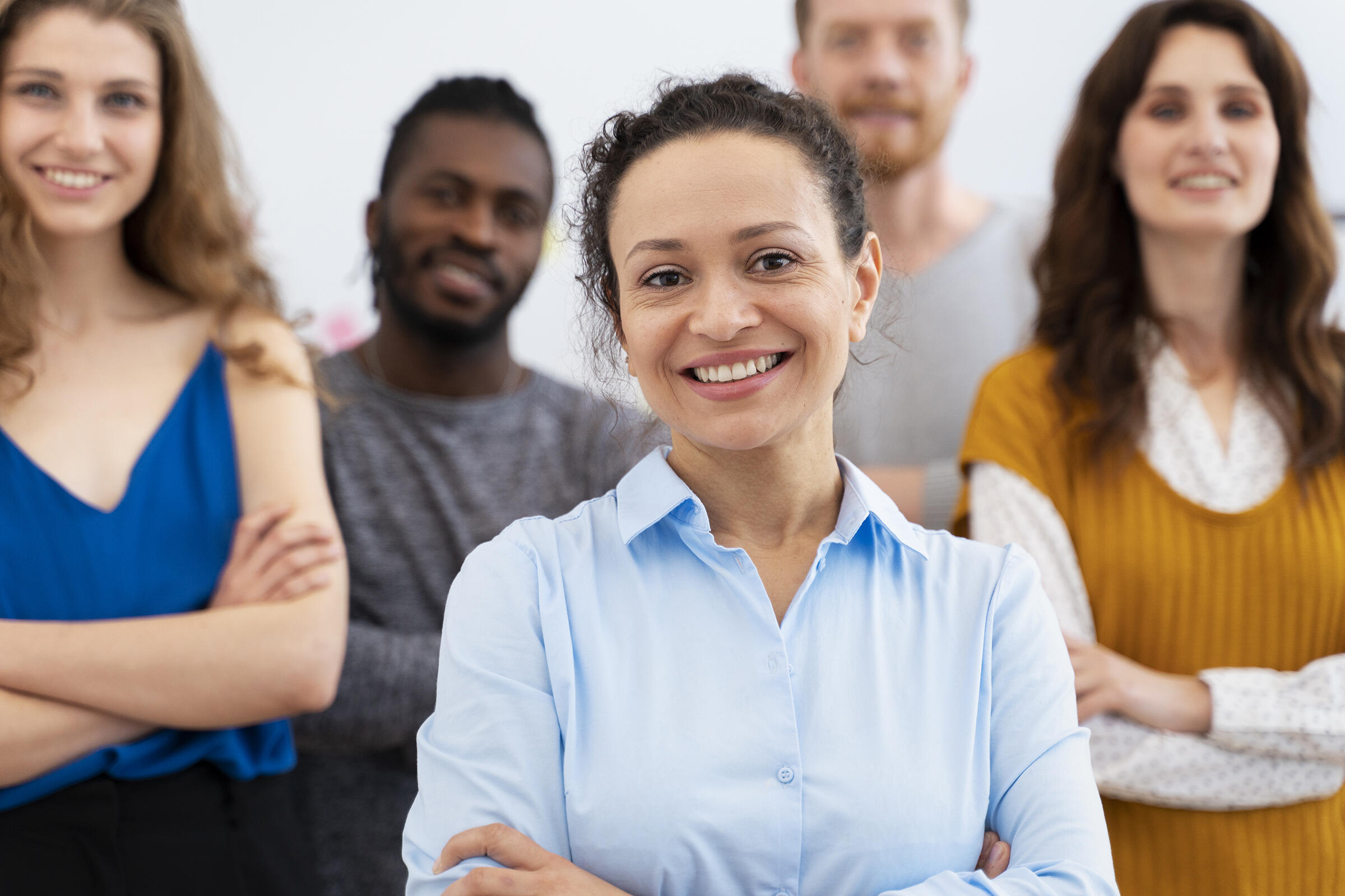 Mehrere Menschen (zwischen 30 und 40), 3 Frauen und 2 Männern, mit unterschiedlicher Hautfarbe schauen lächelnd mit gekreuzten Armen in die Kamera.