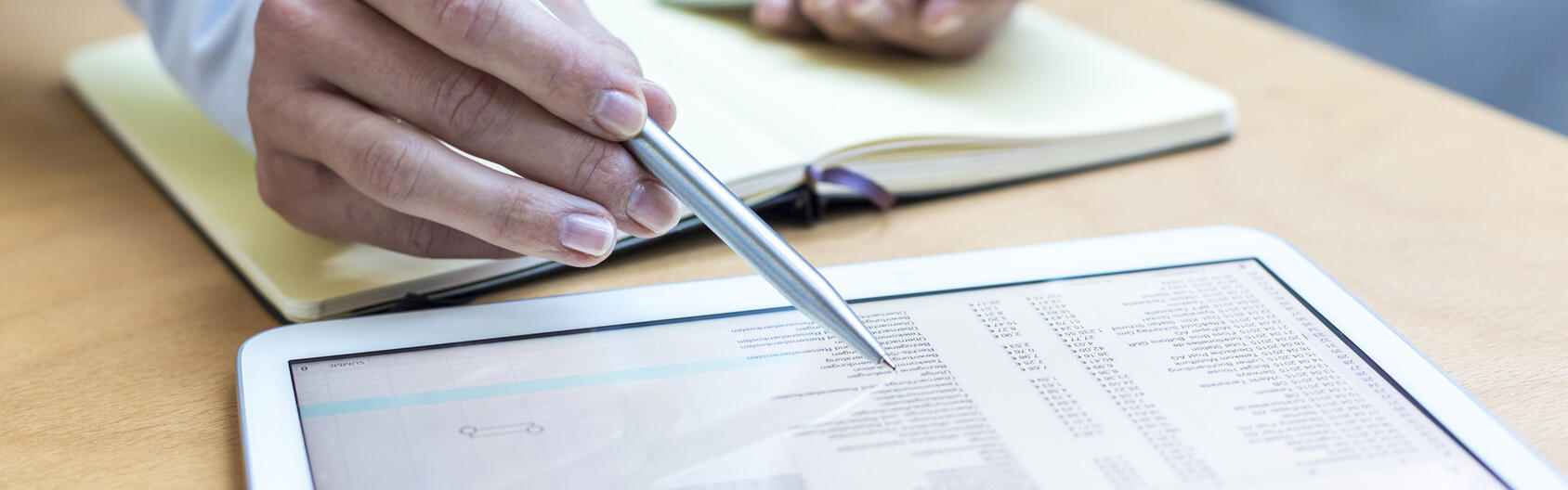 Eine Person im Businessoutfit vergleicht eine Tabelle auf einem Tablet und einem Handy.