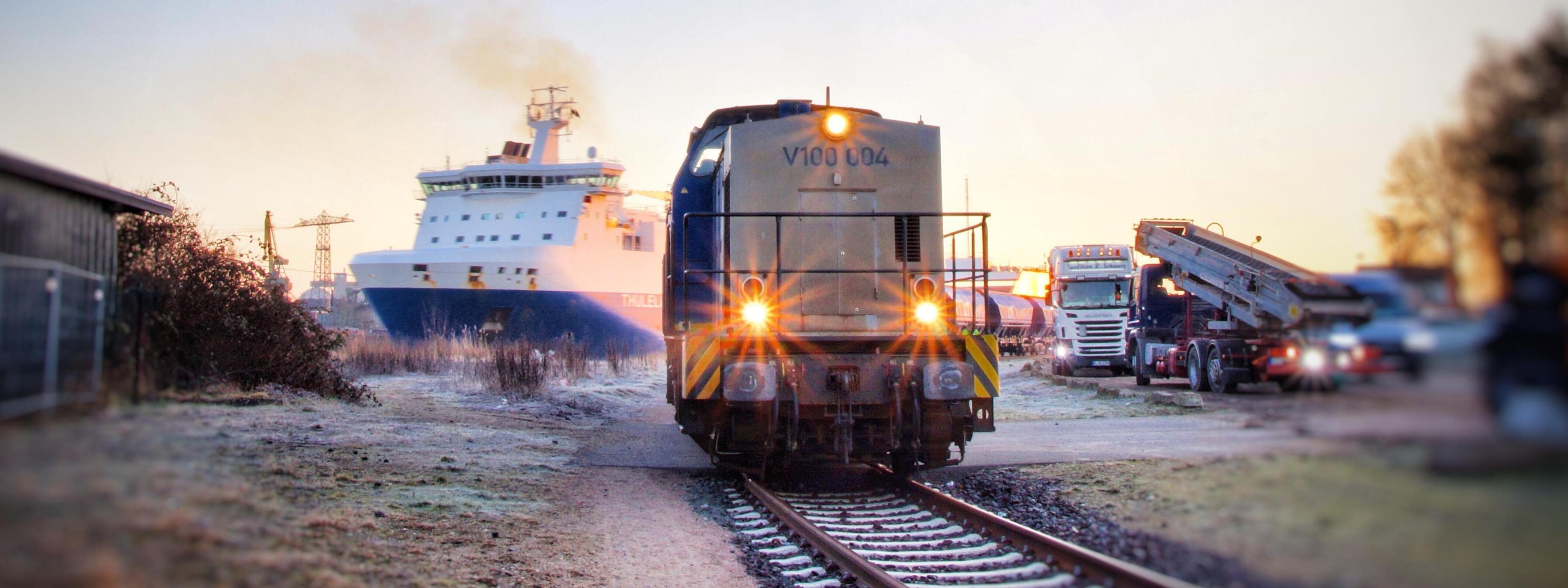 Eine Lok im Vordergrund auf Schienen. Im Hintergrund sieht man ein Schiff, und LKWs