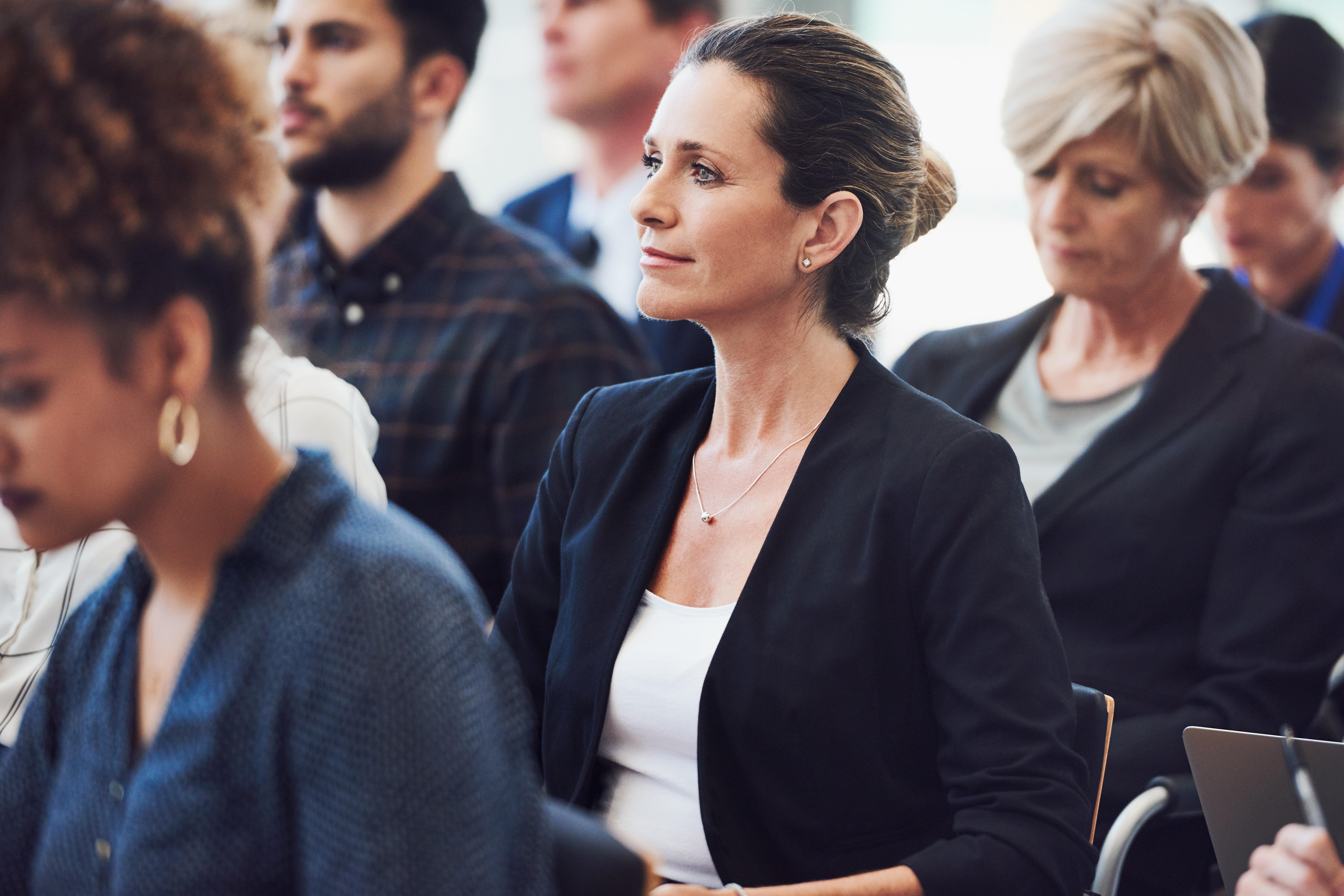 In dem Bild ist sind Teilnehmer einer Konferenz zu sehen, die nach vorne schauen. Im Fokus steht eine Frau, welche inmitten von Personen sitzt die eifrig mitschreiben.
